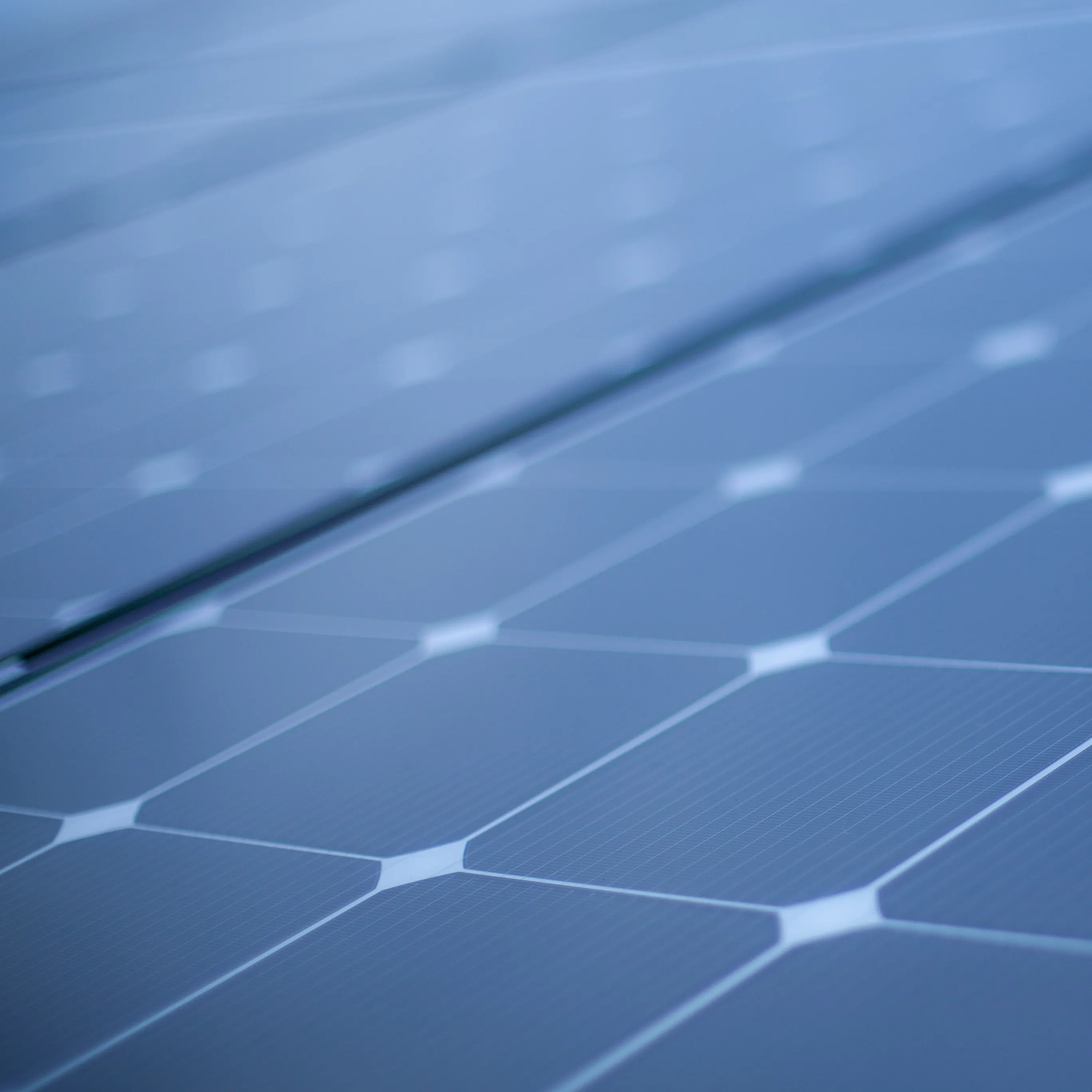Close-up of a solar panel surface, showing its grid-like arrangement of dark photovoltaic cells with white borders. The glossy surface reflects light.
