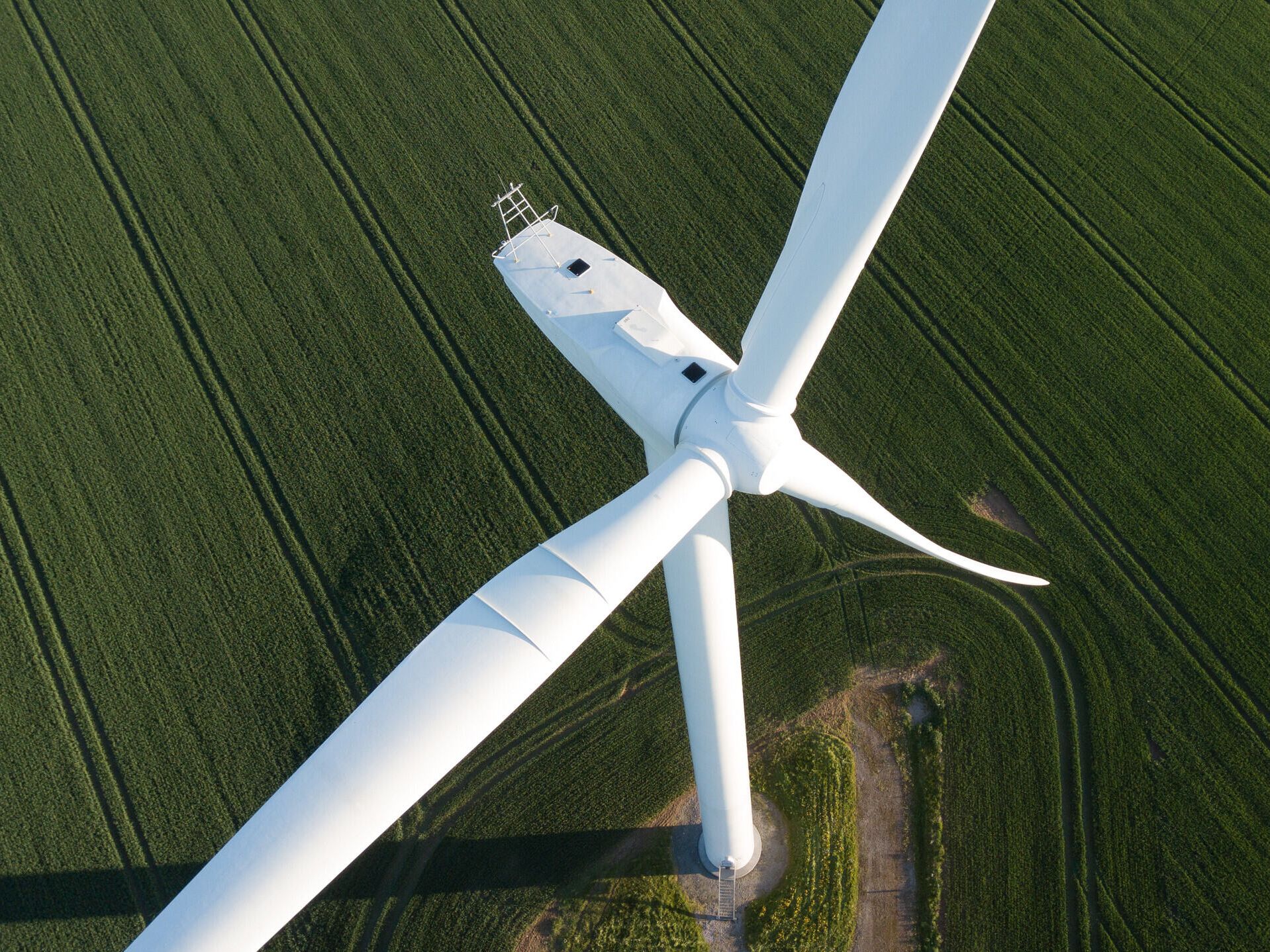 A close-up aerial view of a white wind turbine surrounded by lush green fields. The turbine's blades are captured at an angle, providing a glimpse of its large size. The field is marked with tractor lines, creating a textured landscape.
